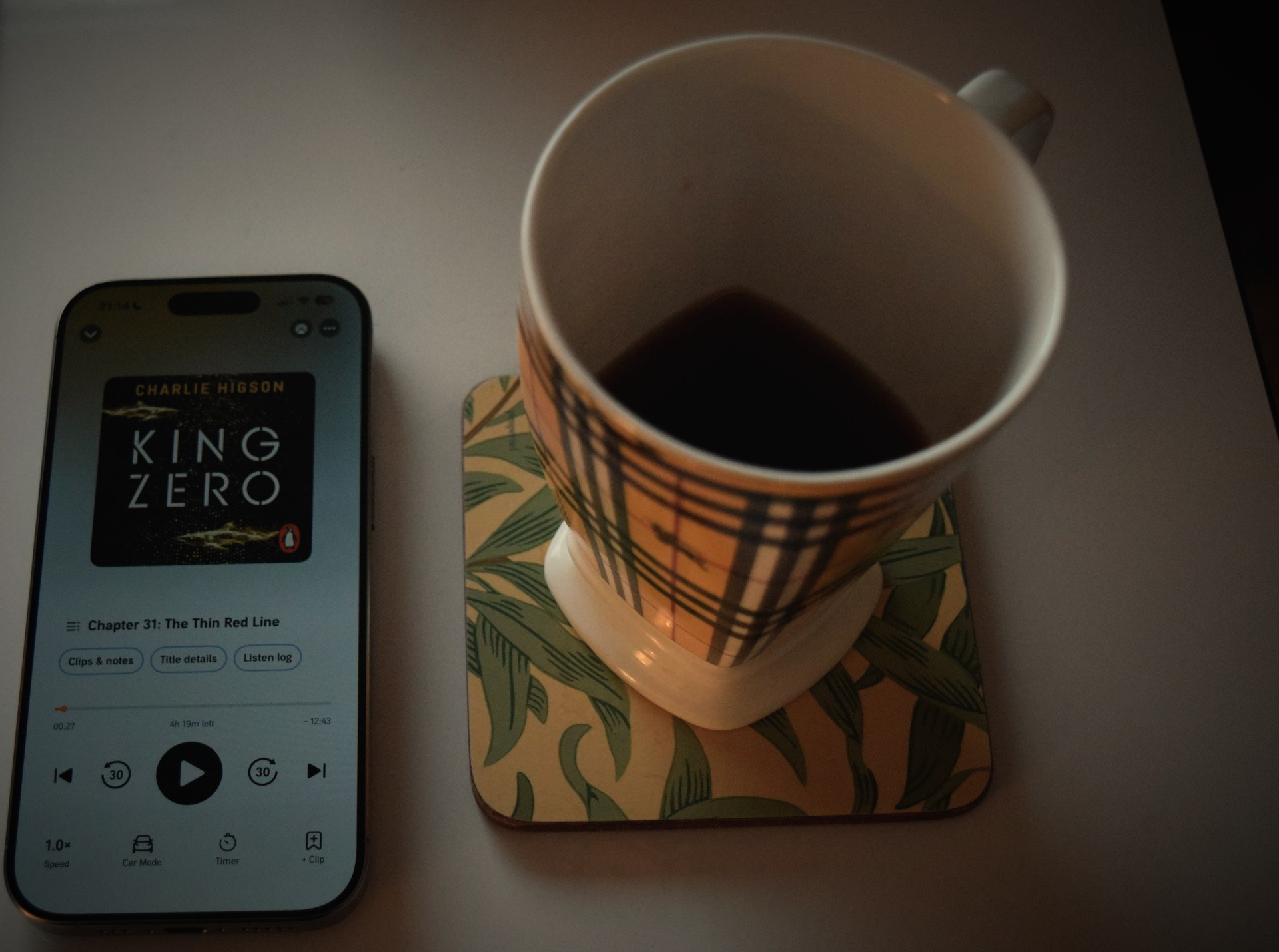 King Zero by Charlie Higson playing on an audiobook app beside a Burberry check mug of cold tea